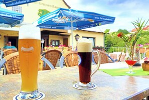 Deux verres à bière sont posés sur une table à l’extérieur, avec un parasol bleu et des chaises en osier en arrière-plan.