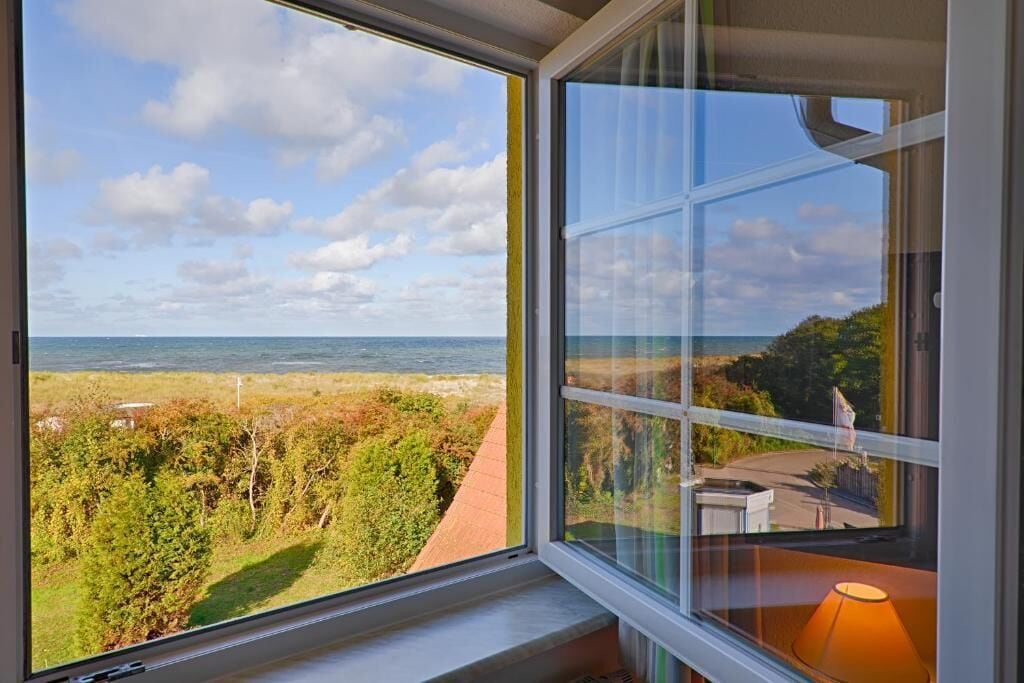 A room with a large window overlooking a coastal landscape, a view of the sea, and a pathway leading to a building.