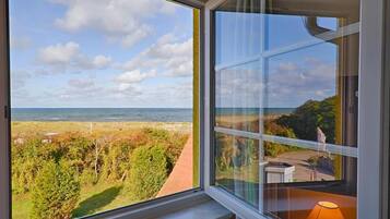 A room with a large window overlooking a coastal landscape, a view of the sea, and a pathway leading to a building.