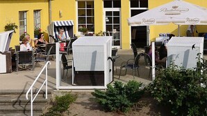 An outdoor seating area with tables and chairs, a yellow building with a red roof, and a white umbrella with 'Pachberger' branding. - Seehotel Düne (Graal Mueritz)
