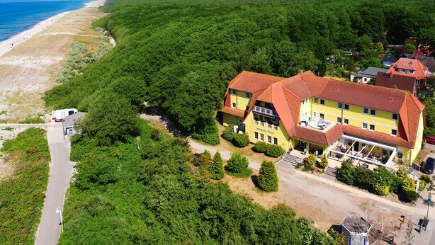 A yellow building with a red roof, surrounded by dense green forest, near a beach. - Seehotel Düne (Graal Mueritz)