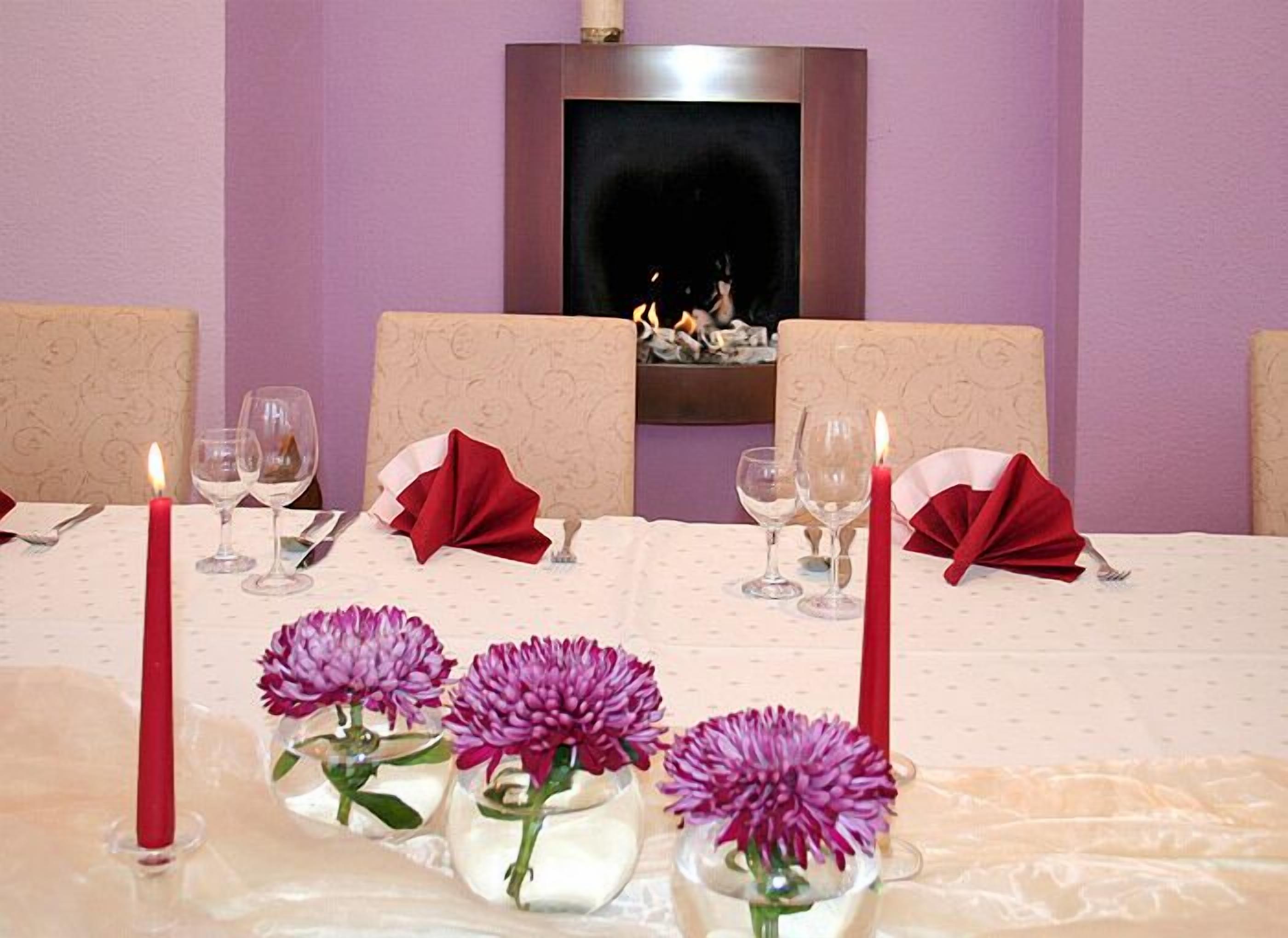 A dining table set for four with red napkins, candles, and purple flowers in glass vases.