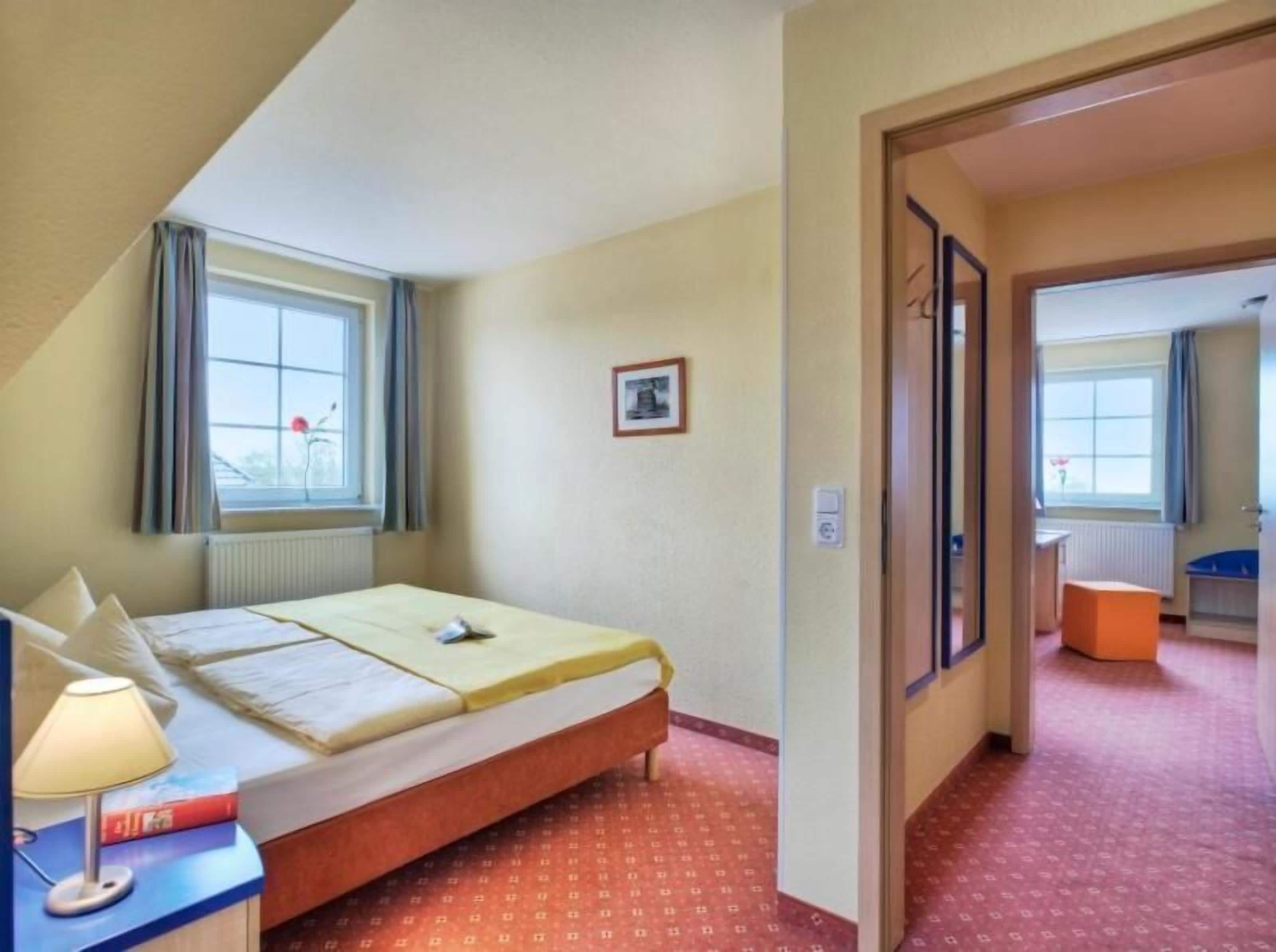 A hotel room with a bed, a desk with a lamp, a window with curtains, and a door leading to another room.