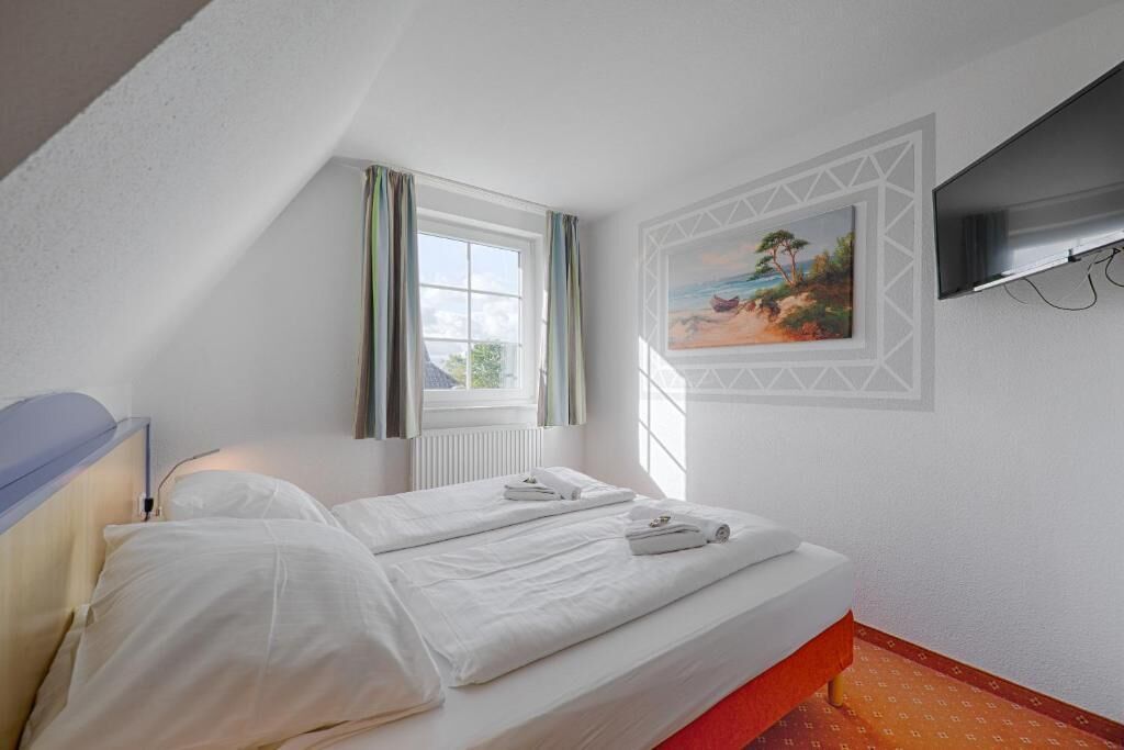 A hotel room with a bed, a window with curtains, a painting on the wall, and a television mounted on the wall.