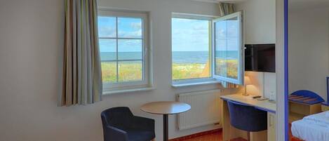 A hotel room with a bed, a desk, a chair, a television, and a window with a view of the beach.