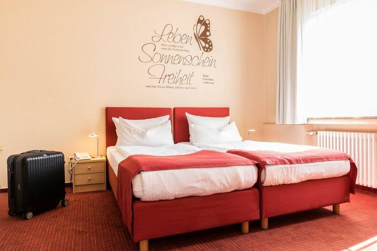 Habitación de hotel con cama roja, una mesita de noche pequeña con lámpara, una maleta y una pared con una mariposa y texto.