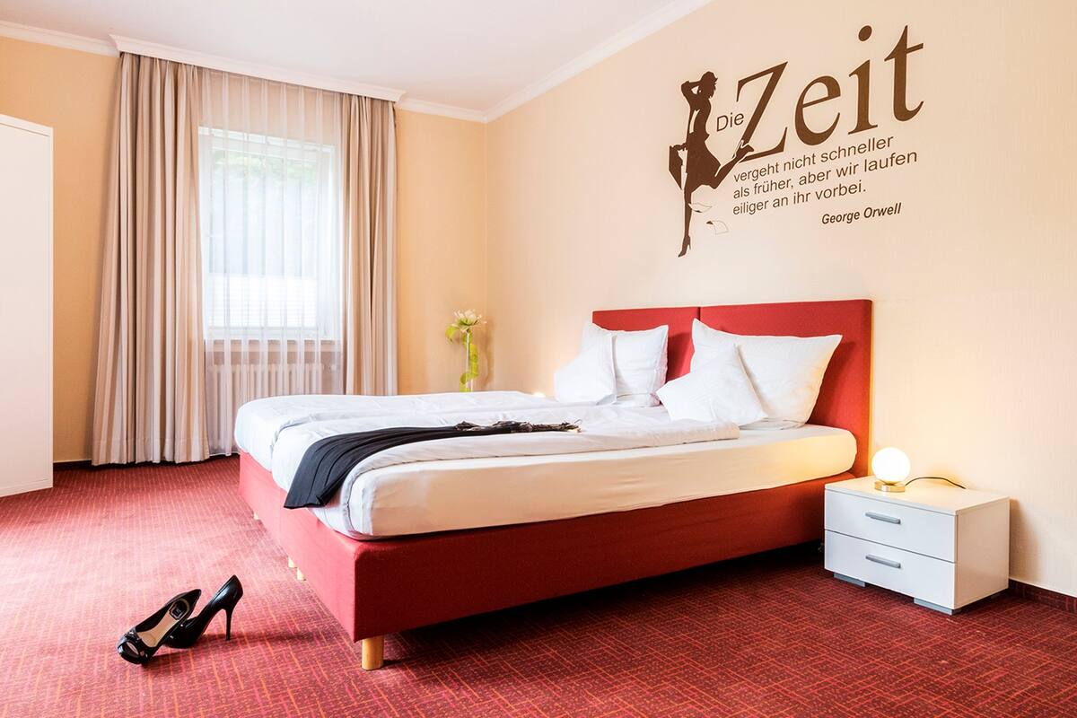 Habitación de hotel con cama roja, almohadas blancas, mesita de noche con lámpara y una cita en la pared.