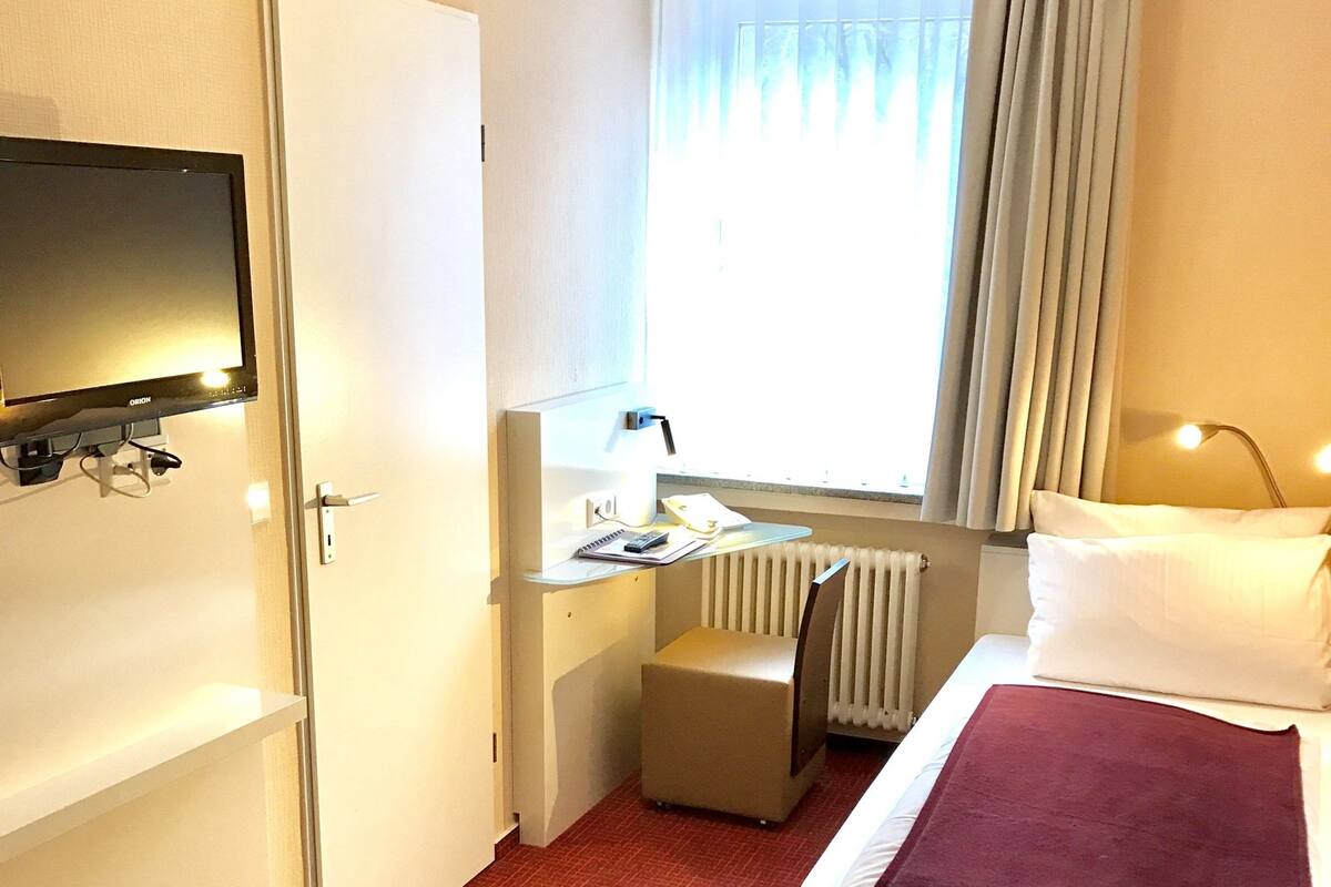 Habitación de hotel con cama, escritorio con computadora, televisor en la pared y ventana con cortinas.
