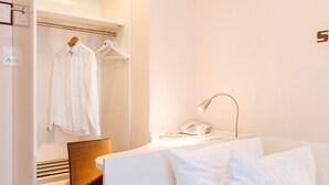 A modern hotel room with a white bed, a brown chair, a white wardrobe, and a bedside table with a lamp and a phone. - Elisabetha (Hannover)