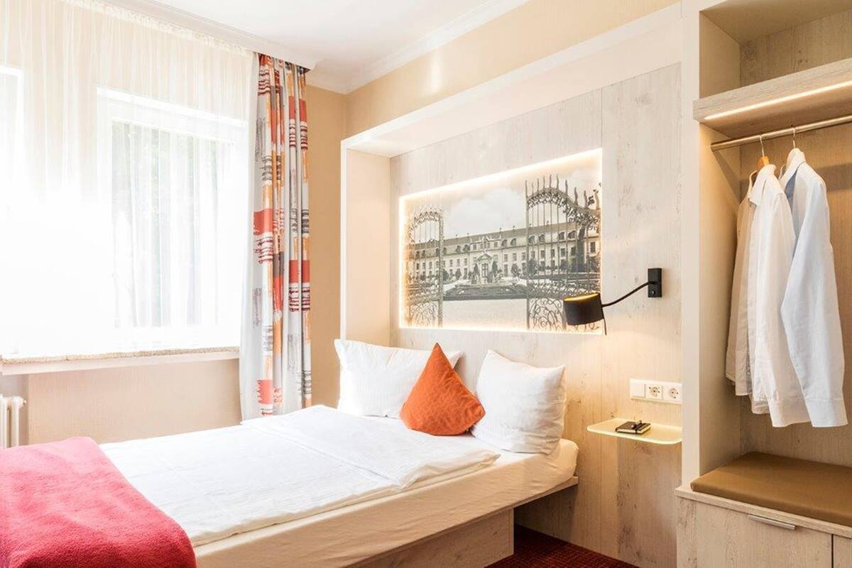 Habitación de hotel con una cama, una mesita de noche, una lámpara de pared y un perchero con ropa.