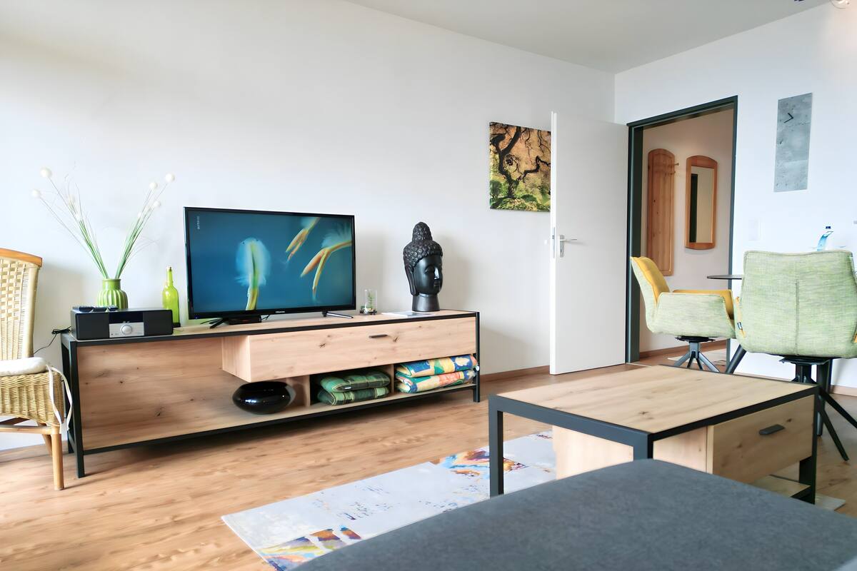 A modern living room with a wooden floor, a flat-screen TV on a wooden stand, a green chair, a small table, and a wall-mounted artwork.