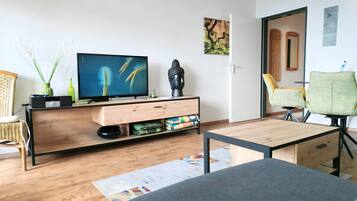 A modern living room with a wooden floor, a flat-screen TV on a wooden stand, a green chair, a small table, and a wall-mounted artwork.