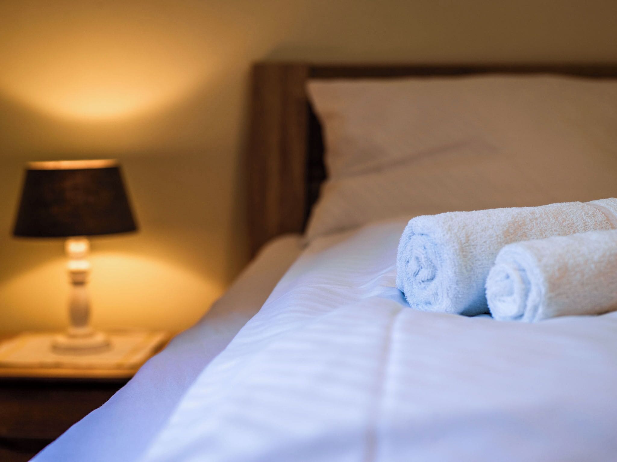 Una cama bien hecha con sábanas blancas y toallas enrolladas, una lámpara de noche y un cabecero de madera.