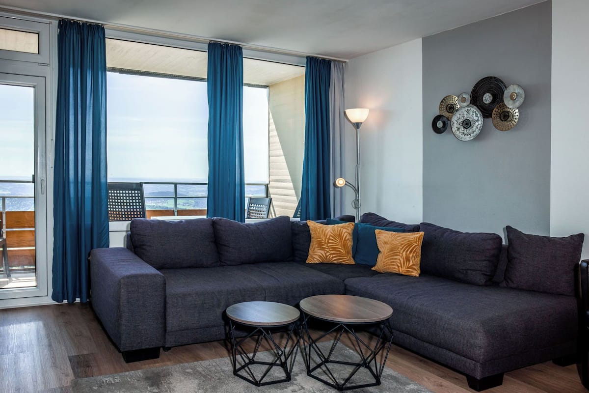 A modern living room with a sectional sofa, two round tables, and a view of the sea through large windows.
