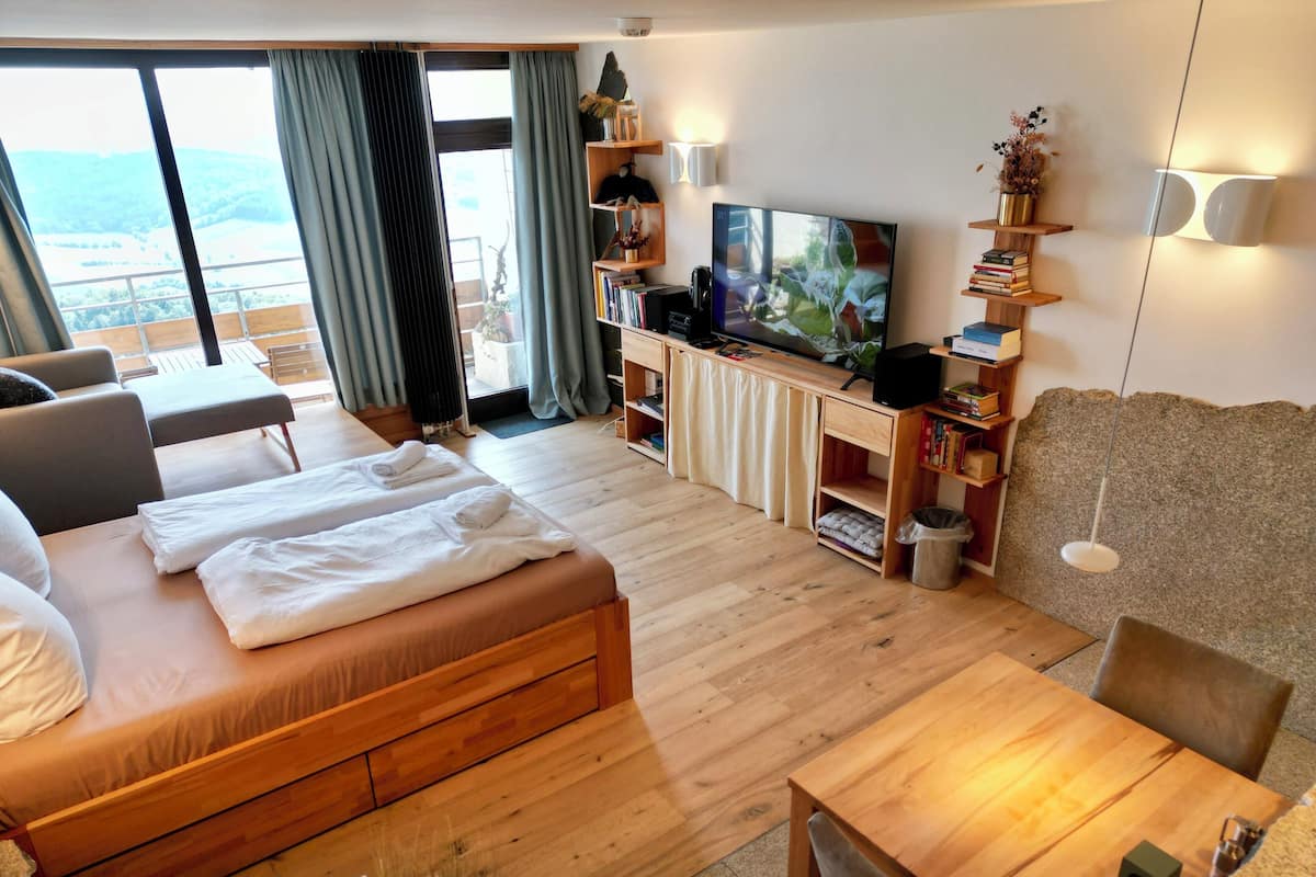 A modern hotel room with a large bed, a TV, a wooden desk, and a balcony with a view.