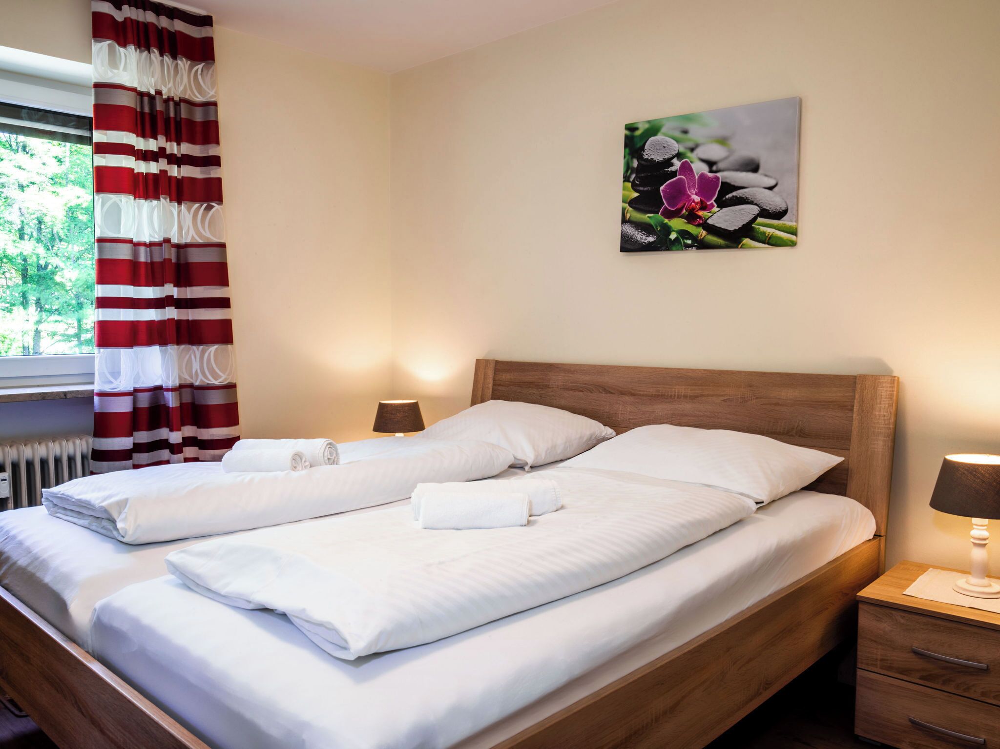 Ein Doppelbett mit weißer Bettwäsche, ein Holz-Kopfstück, ein Nachttisch mit Lampe und ein Blumenbild an der Wand.