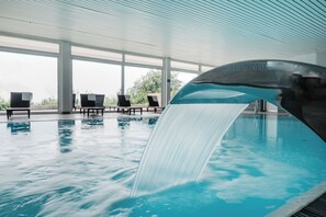 An indoor swimming pool with a waterfall feature, lounge chairs, and large windows. - Ferienpark Geyersberg inklusive aktivCARD Bayerischer Wald (Freyung)