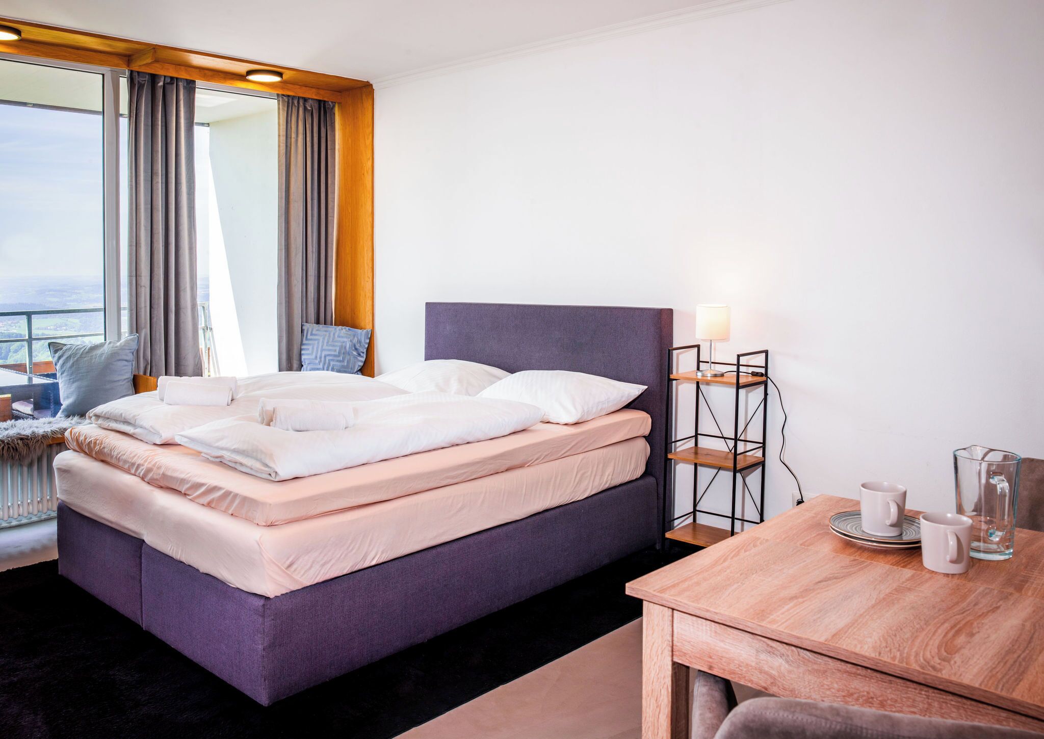 Habitación de hotel con cama, mesita de noche con lámpara, vista a la ciudad y ventana con cortinas.