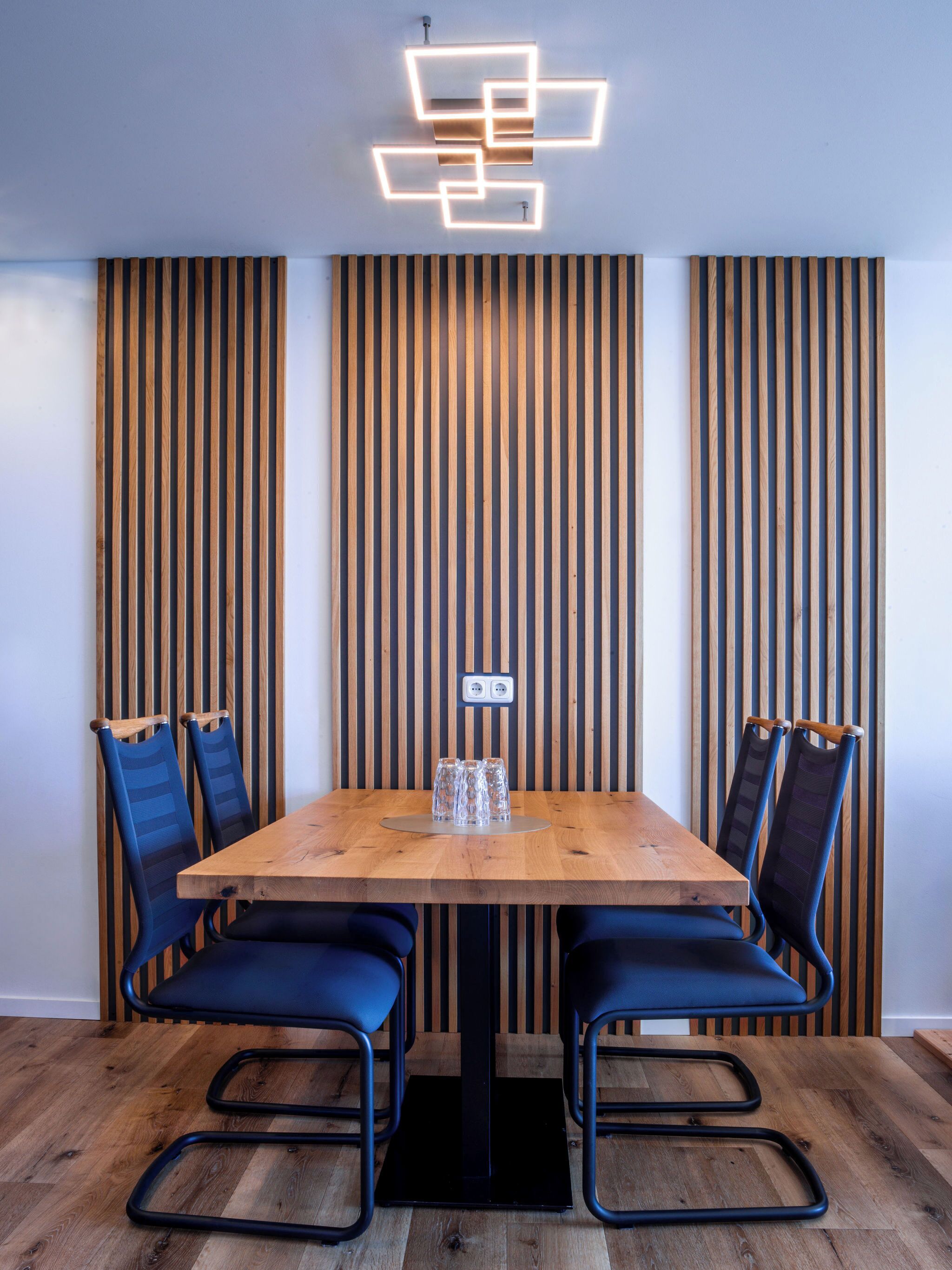A wooden dining table with four chairs, a modern light fixture, and a vertical wooden panel wall.