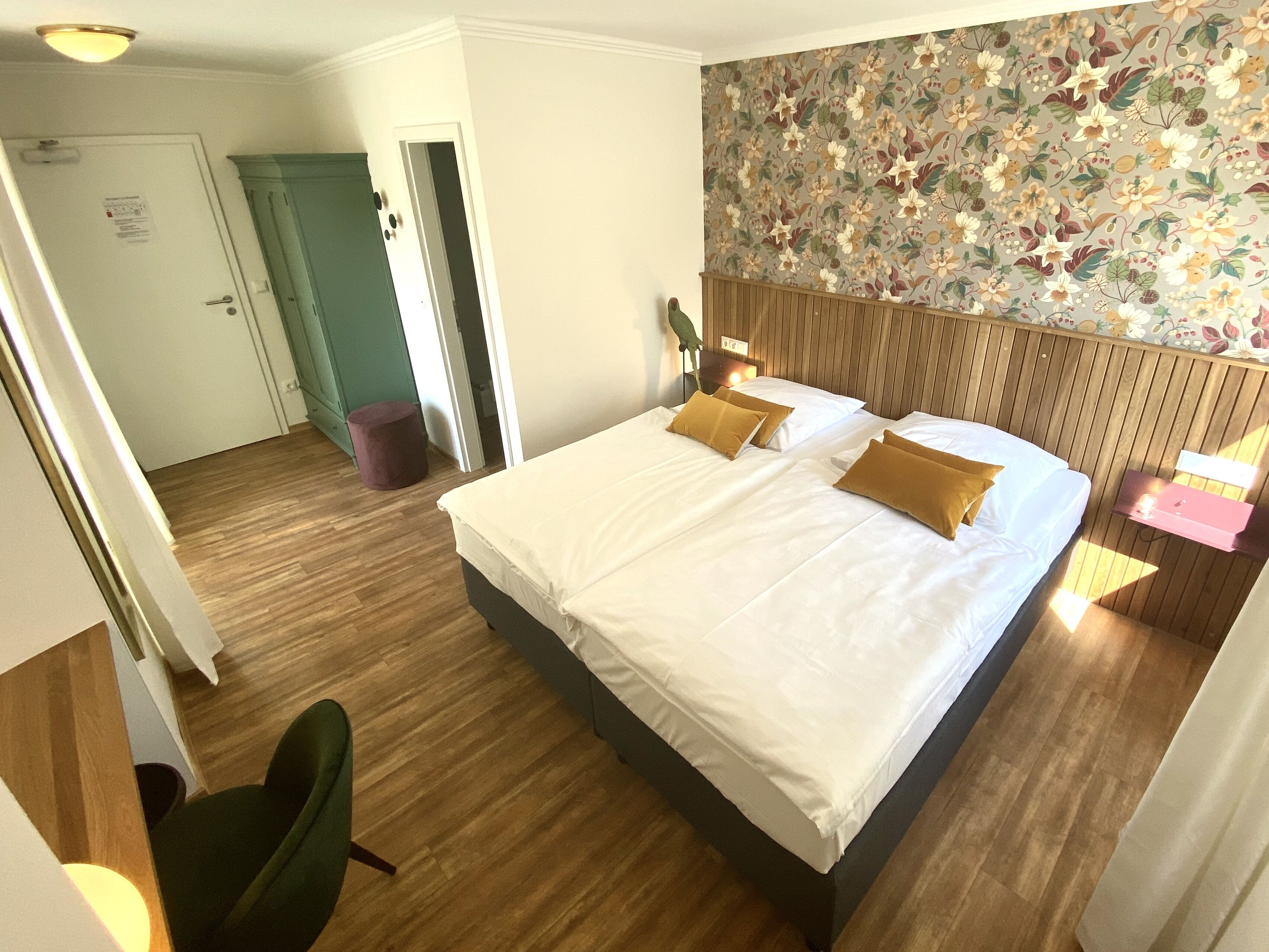 A hotel room with a large bed, wooden flooring, a green chair, a wooden headboard, and a floral wallpaper.