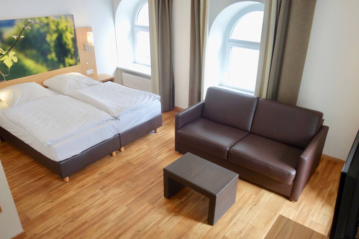 A hotel room with a bed, a brown leather sofa, a small table, and large windows with curtains.