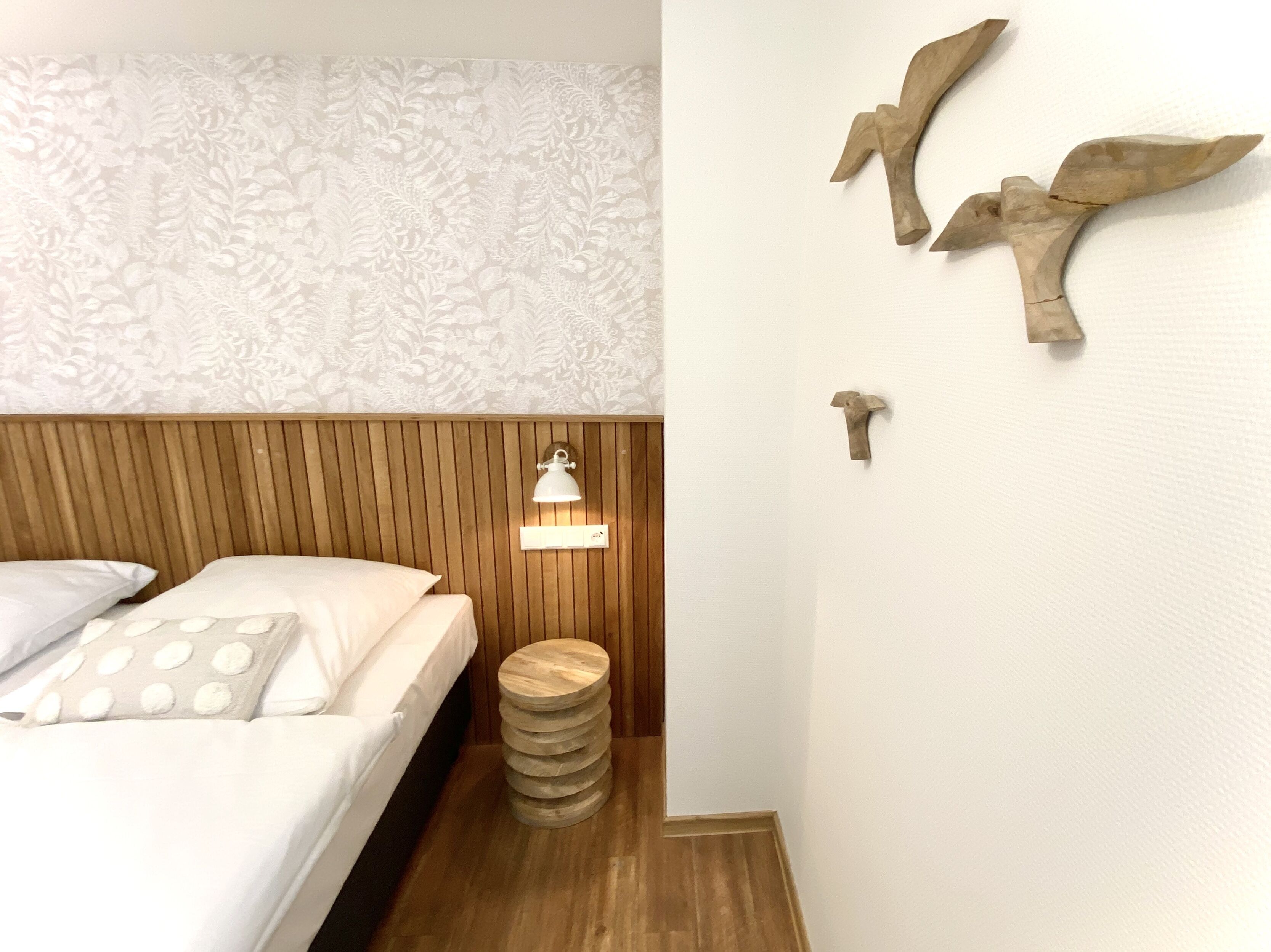 Quarto com cabeceira de madeira, cama com roupa de cama branca, um banquinho de madeira redondo, luminária de parede e decoração de pássaros na parede.