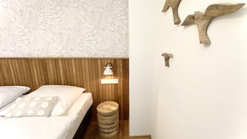 Quarto com cabeceira de madeira, cama com roupa de cama branca, um banquinho de madeira redondo, luminária de parede e decoração de pássaros na parede.