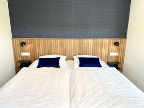 Cama doble con sábanas blancas y dos almohadas azul oscuro, cabecera de madera panelada, dos lámparas de noche y una pared con diseño.