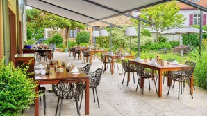 Un espace de restauration en plein air, avec plusieurs tables et chaises, entouré de verdure et agrémenté d’un bâtiment en pierre en arrière-plan.