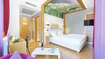A modern hotel room with a bed, a desk with a chair, a small table with a magazine, and a TV on the wall.