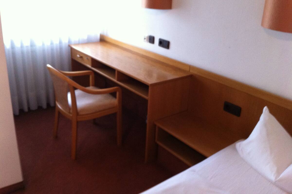 A hotel room with a wooden desk, a chair, a bed with white linens, and a wall-mounted lamp.