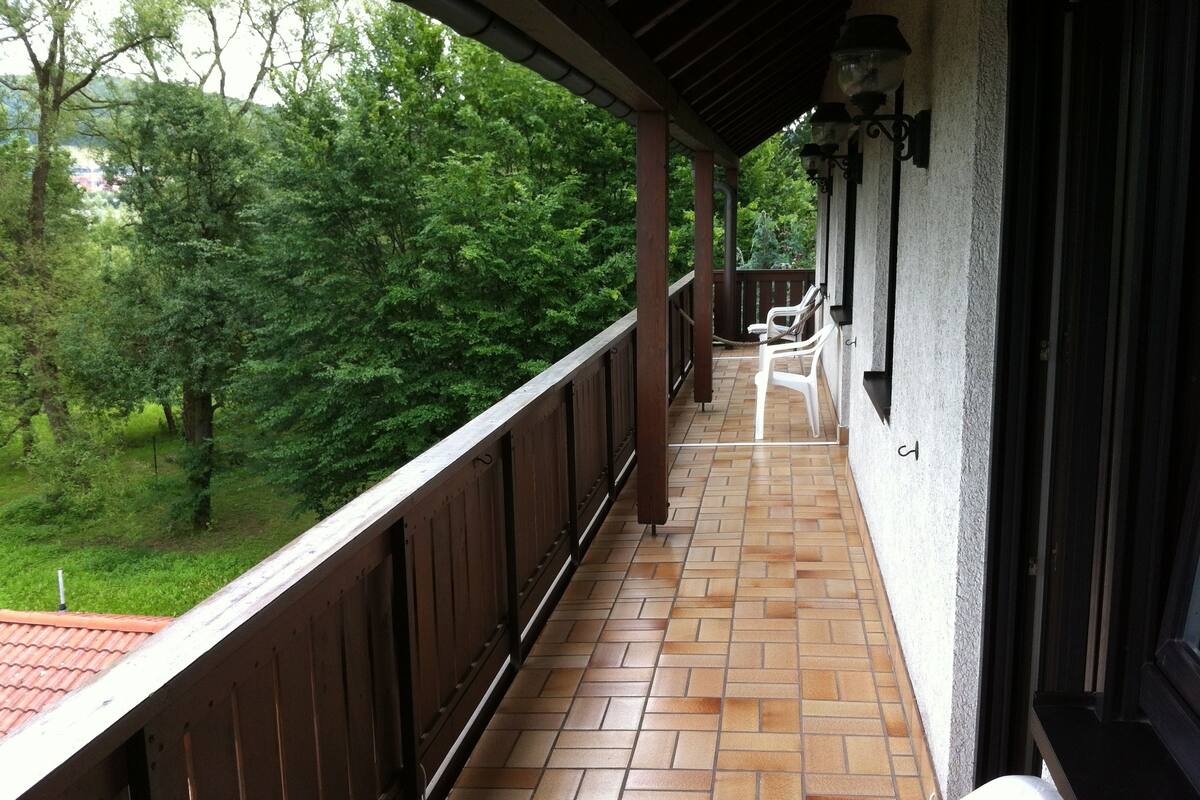 A covered balcony with a tiled floor, wooden railing, and two white plastic chairs.