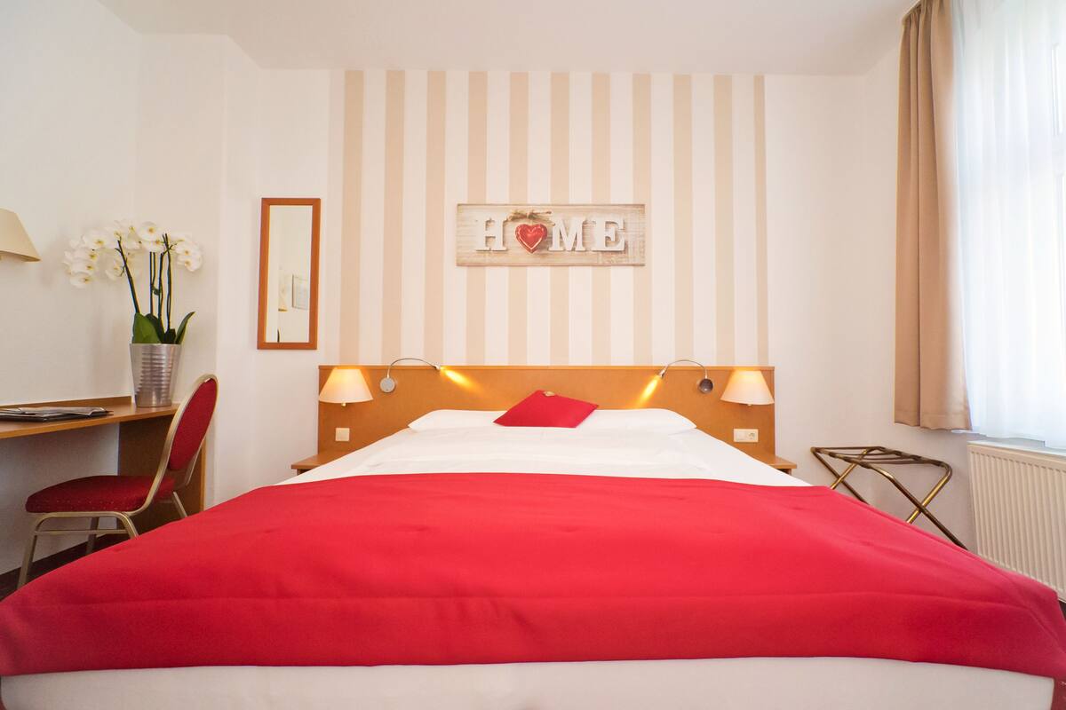 A hotel room with a bed, a desk, a chair, a lamp, a mirror, and a sign that says "HOME".