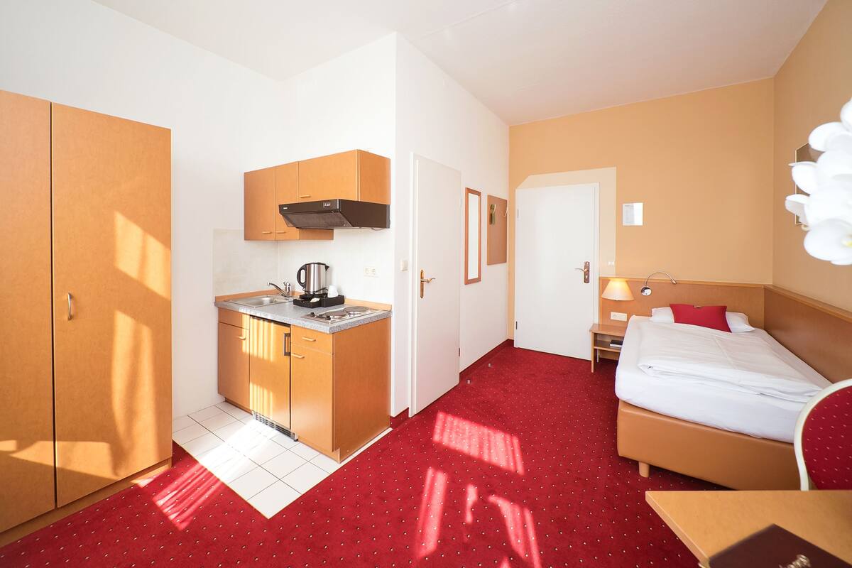 A compact hotel room with a kitchenette, a bed, and a red carpet.