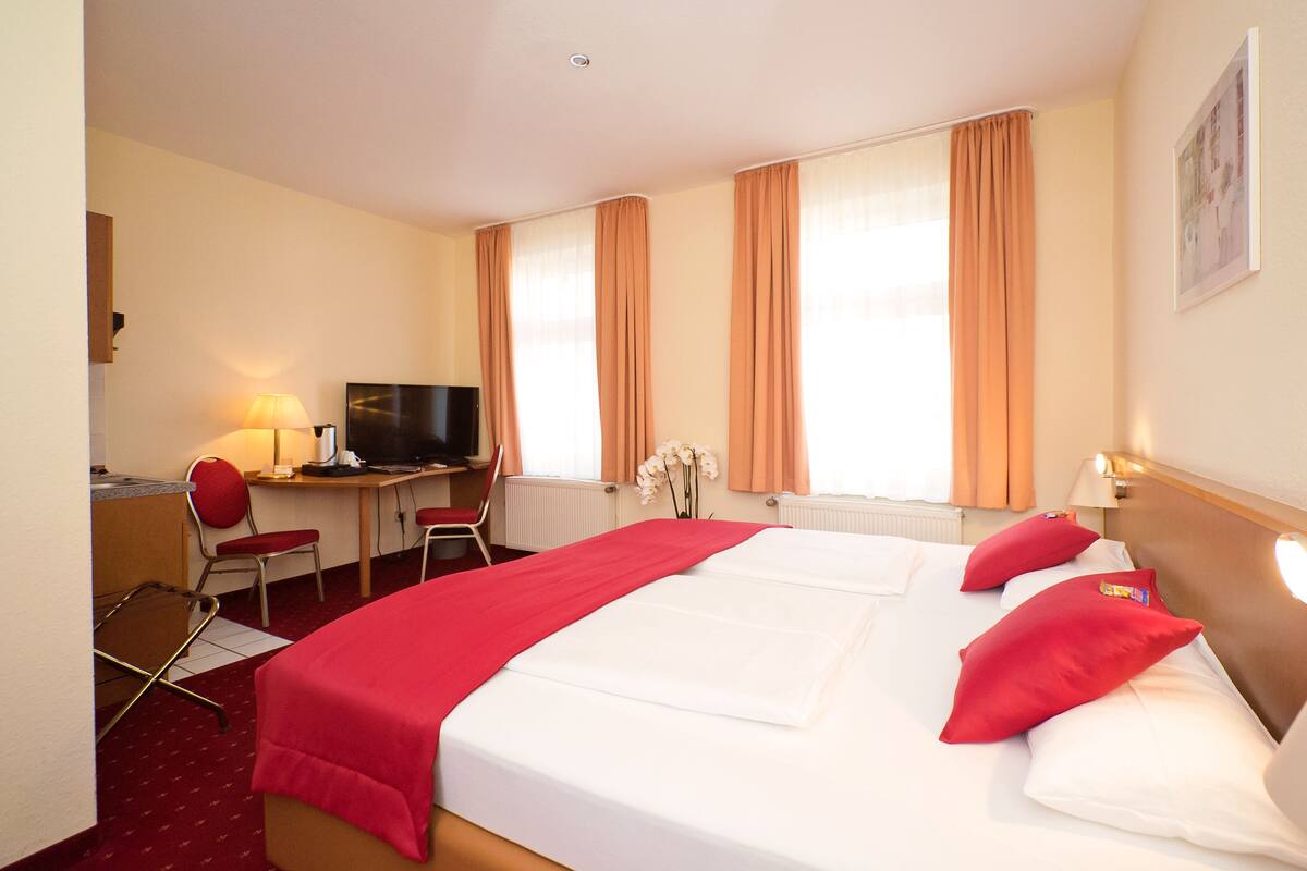 A hotel room with a bed, a desk with a television, a chair, and a window with curtains.