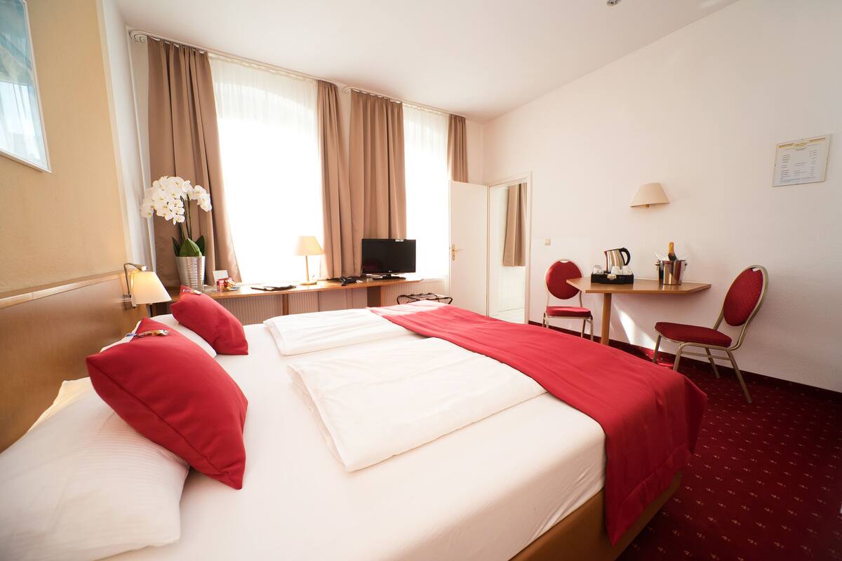 A hotel room with a large bed, a desk with a television, a chair, and a small table with a lamp and a coffee pot.