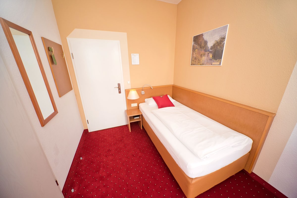 A hotel room with a single bed, a nightstand, a lamp, a door, and a painting on the wall.