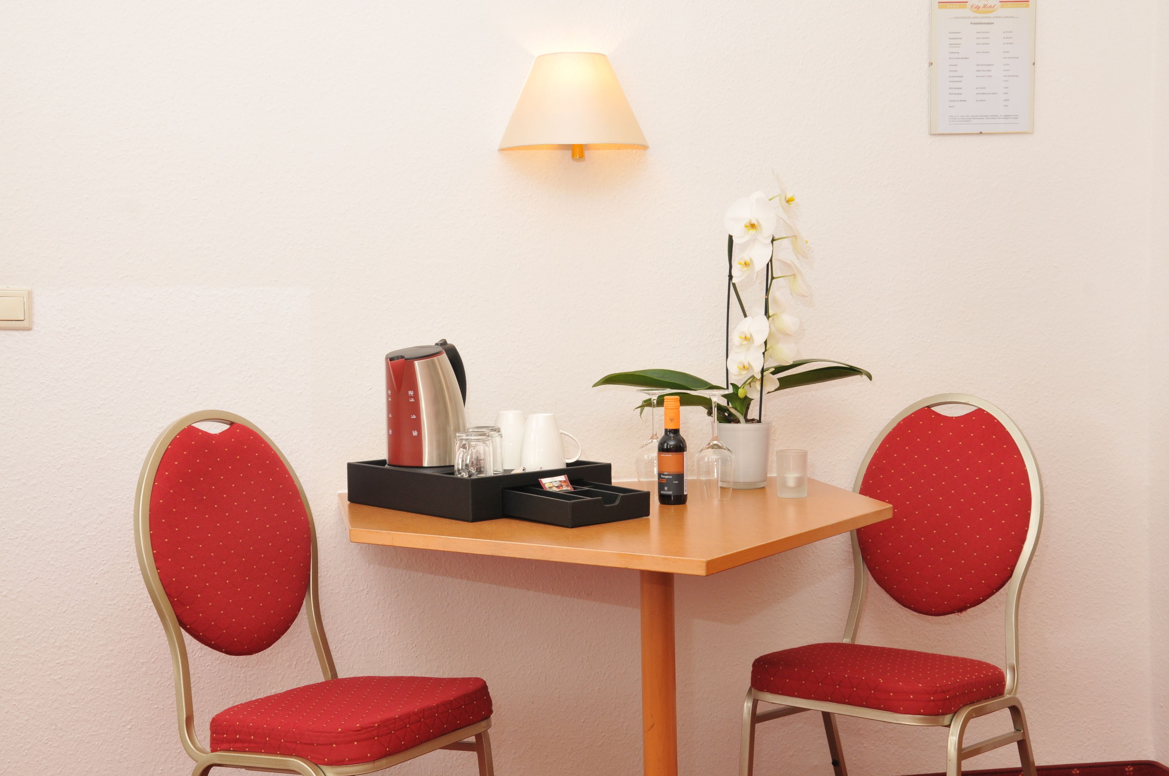 Habitación de hotel con una mesa de madera, dos sillones tapizados en rojo, una lámpara, una orquídea en maceta y un minibar.
