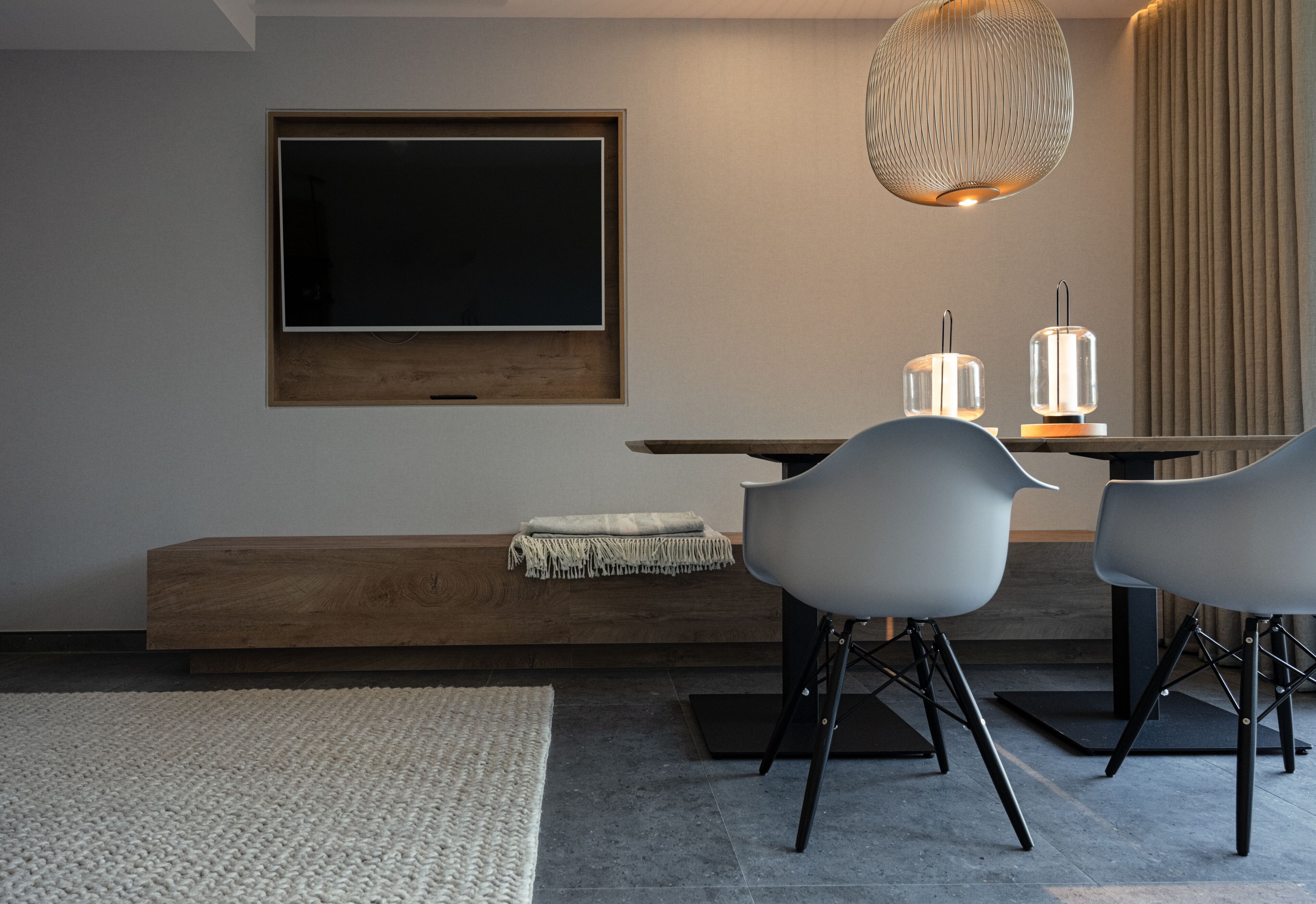 Una sala moderna con un televisor de pantalla plana grande montado en la pared, un banco de madera y una mesa de comedor con dos sillas.