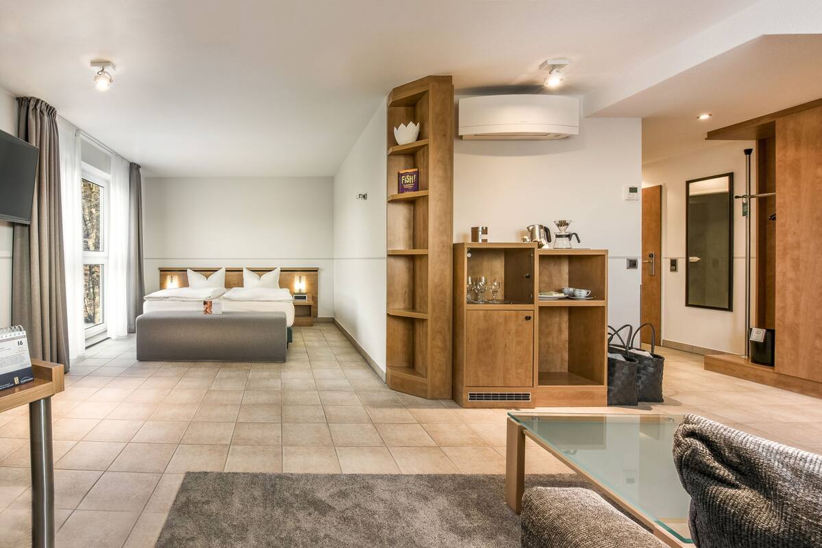 Una habitación de hotel moderna con una cama grande, una mesa de centro de vidrio, un sofá, un armario de madera y un guardarropa.