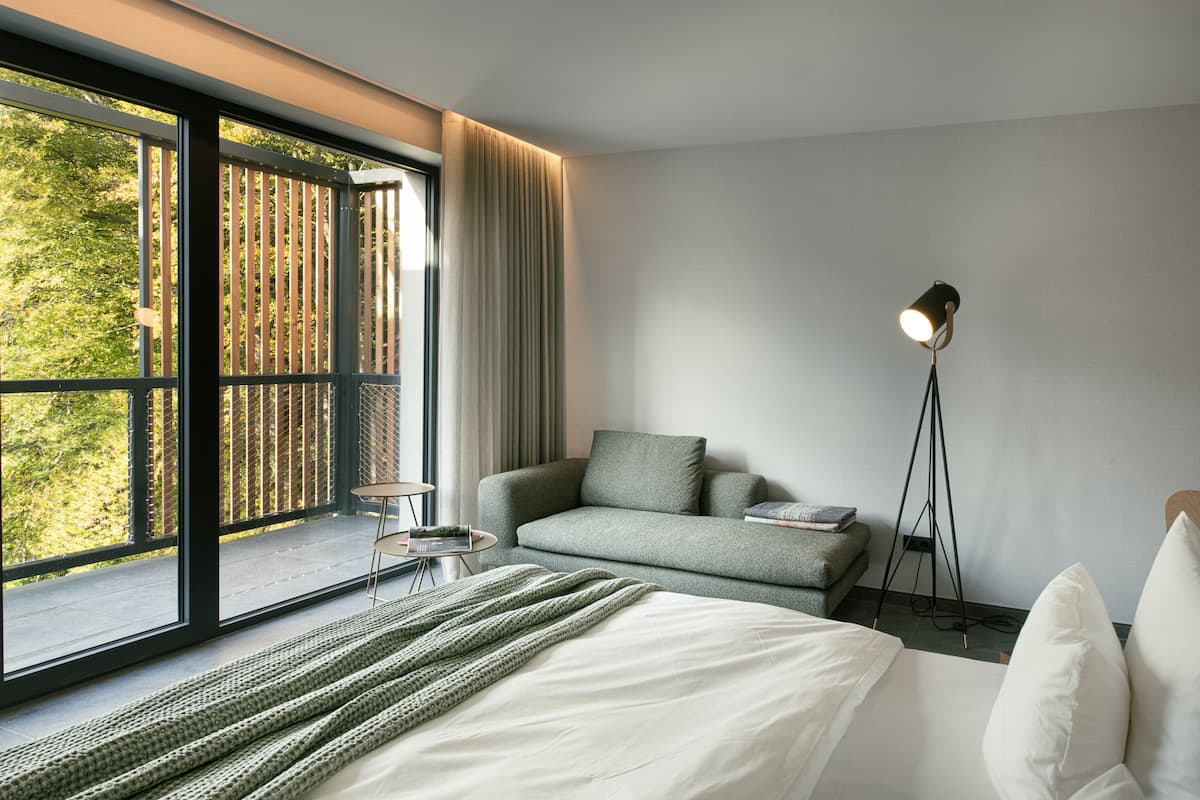 Una habitación de hotel moderna con un ventanal grande, un sofá, una mesita de noche y una lámpara de pie.