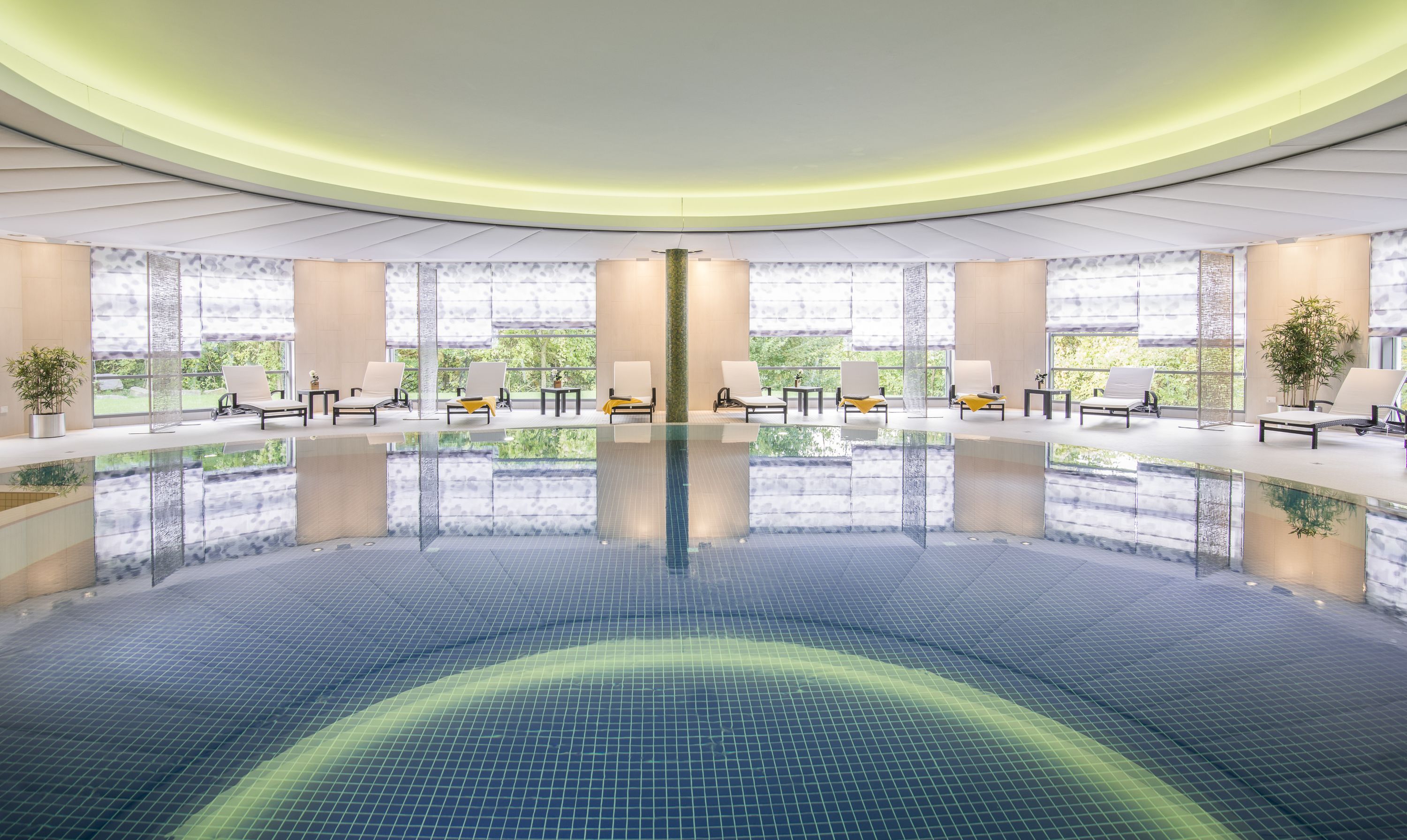 Una amplia piscina interior con sillones de descanso y mesas, rodeada de grandes ventanales y un moderno diseño de techo.