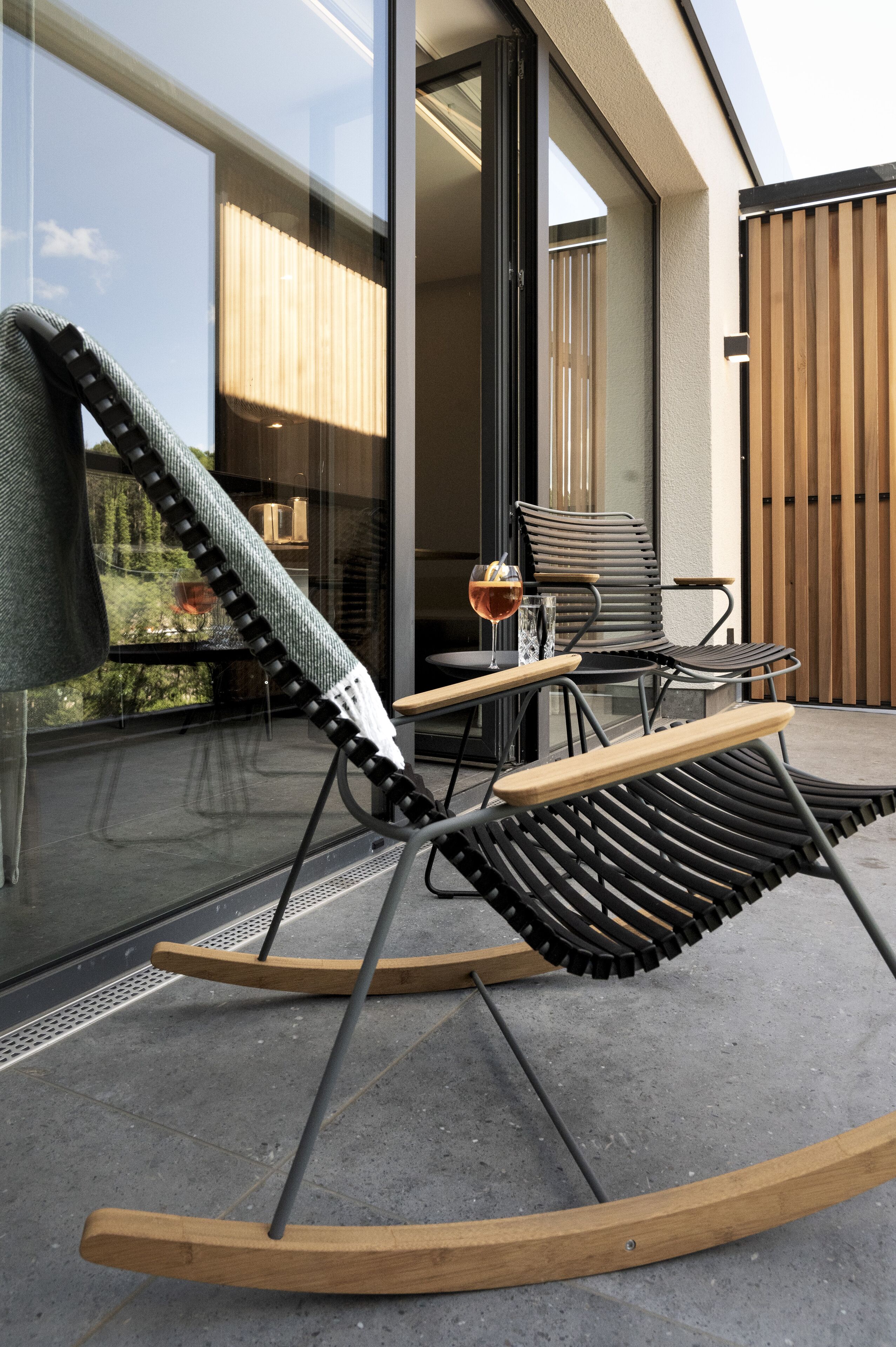A modern outdoor seating area with a rocking chair and a table, a glass of drink, and a view of the interior through a glass door.