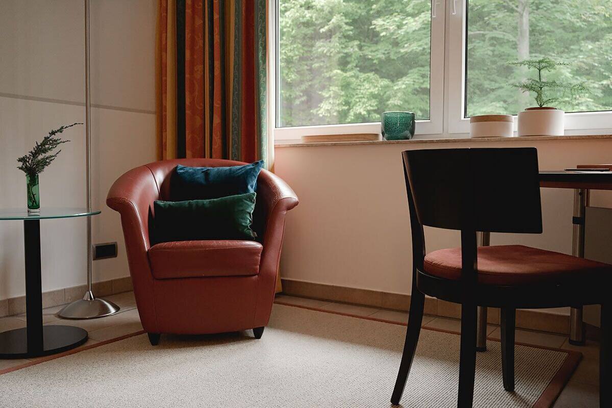 Una habitación con un sillón de cuero marrón, una silla negra, una mesa de vidrio y vistas a árboles a través de una ventana.