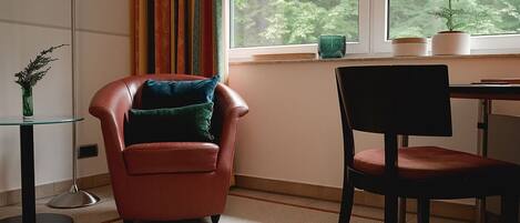 Una habitación con un sillón de cuero marrón, una silla negra, una mesa de vidrio y vistas a árboles a través de una ventana.