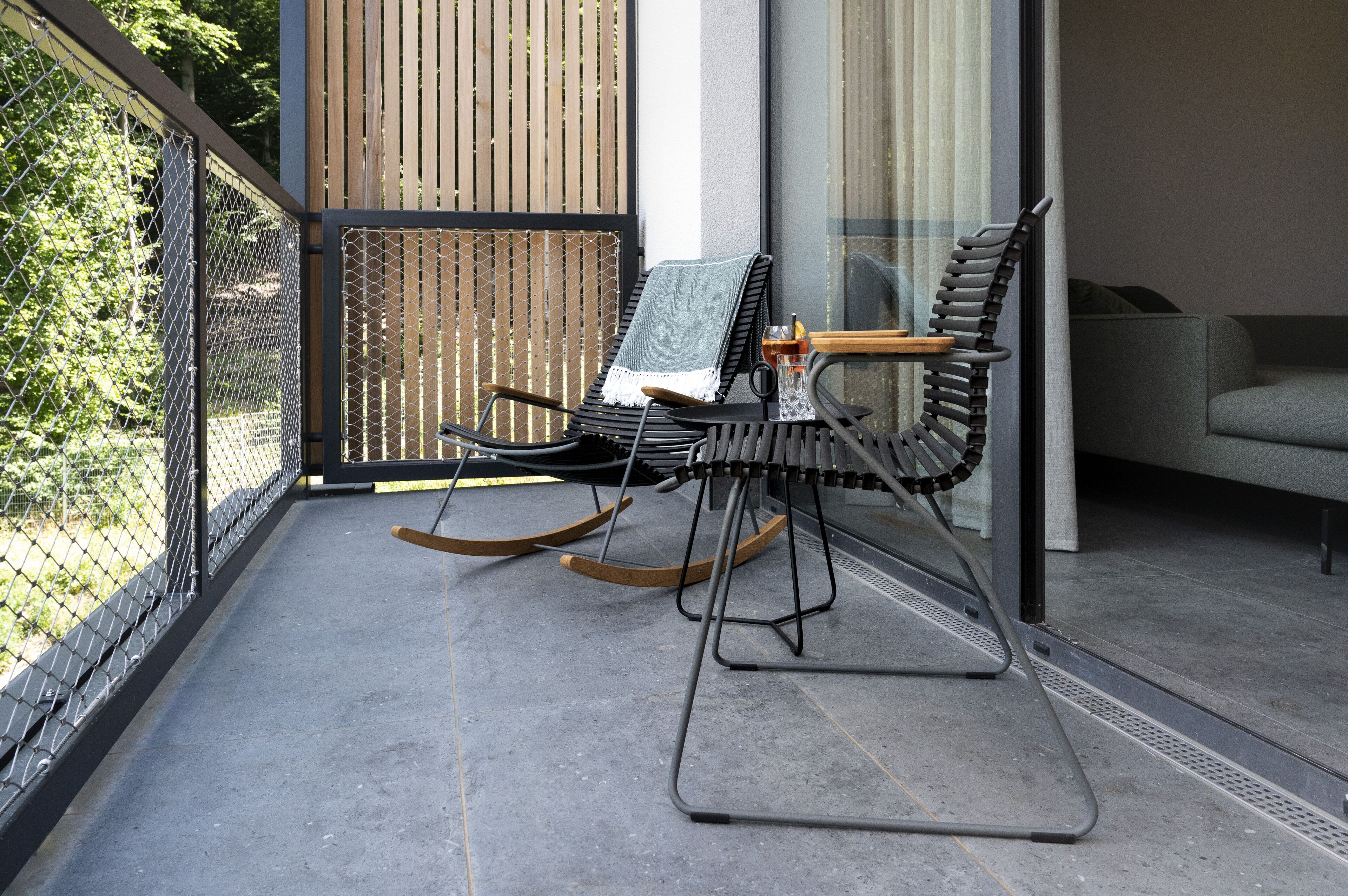 A balcony with two rocking chairs, a glass table, and a view of trees.