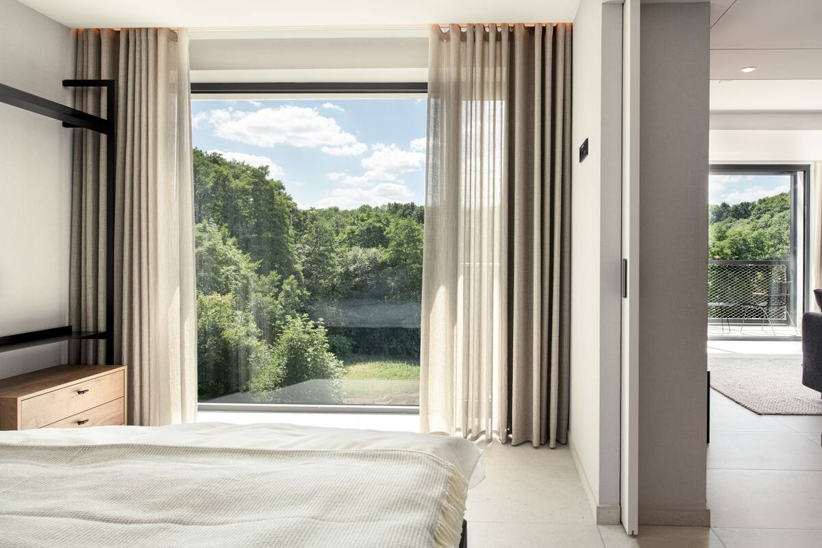 Un dormitorio con una ventana grande, una cama, una mesita de noche y vistas a un bosque.
