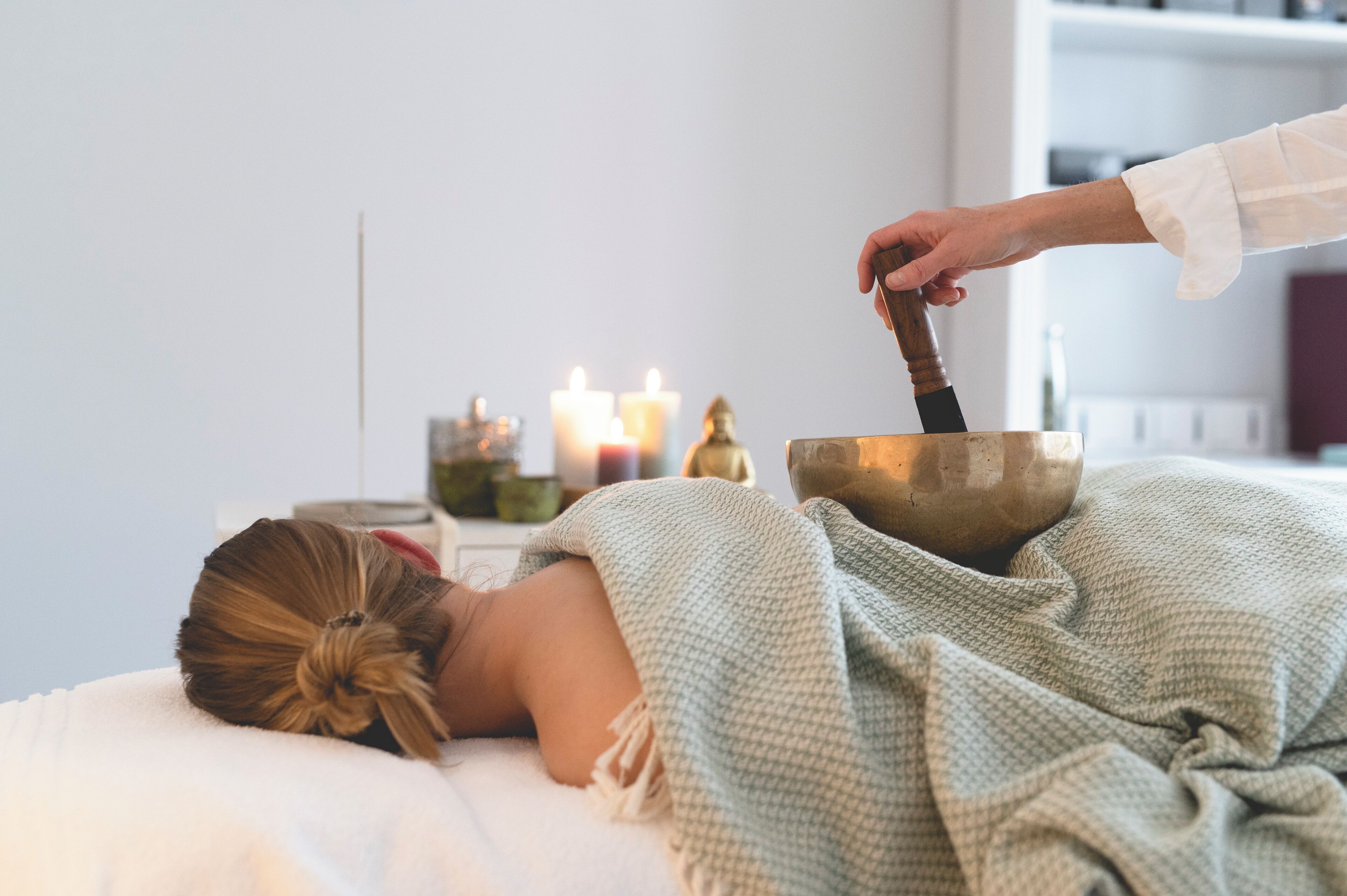 A person lying on a bed with a blanket, a bowl, and a mallet, suggesting a meditation or relaxation setting.