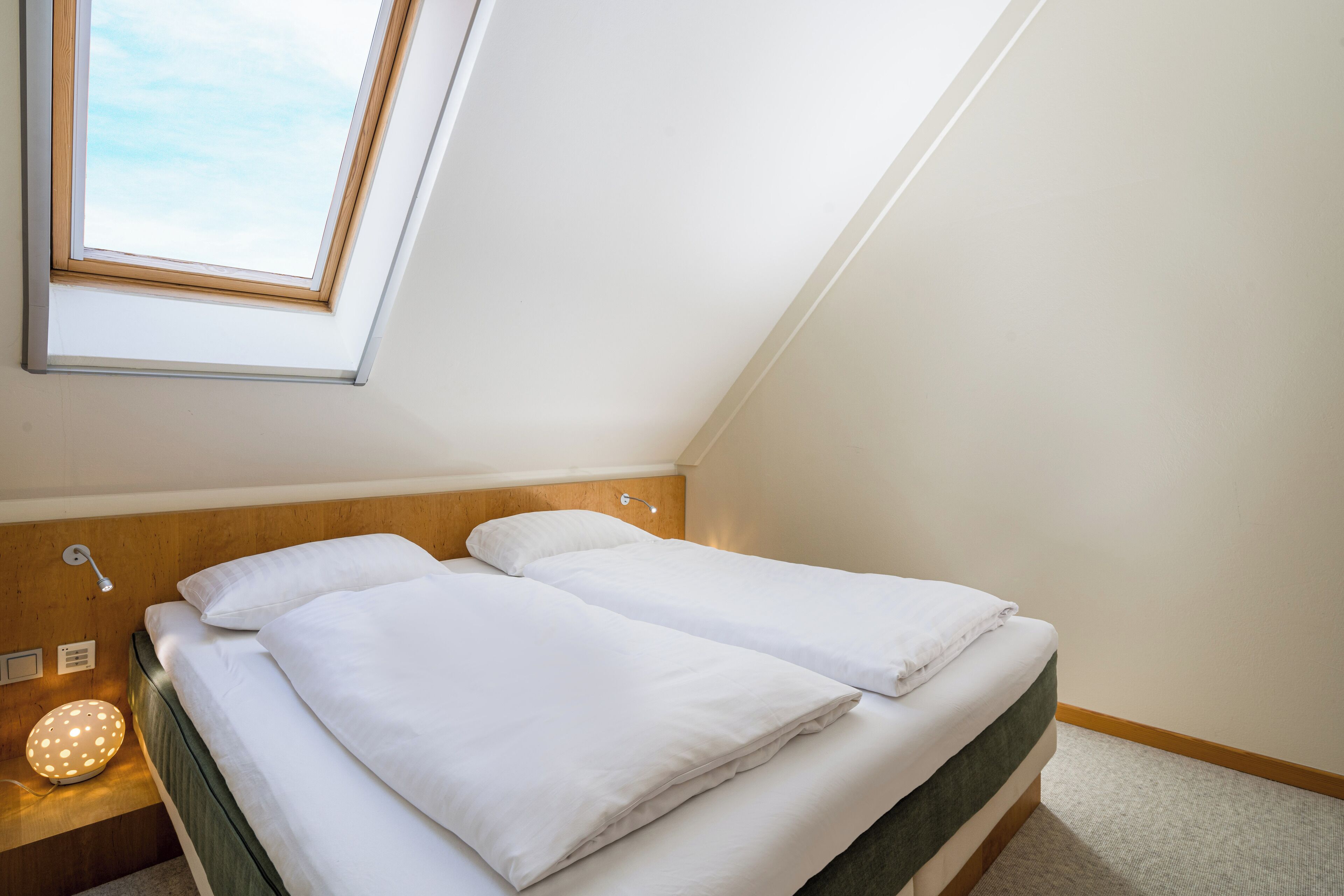 Ein Doppelbett mit weißer Bettwäsche in einem Zimmer mit schrägem Dach und Dachfenster.