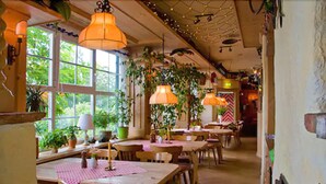 Un restaurante acogedor con mesas y sillas de madera, ventanas amplias y plantas colgantes.